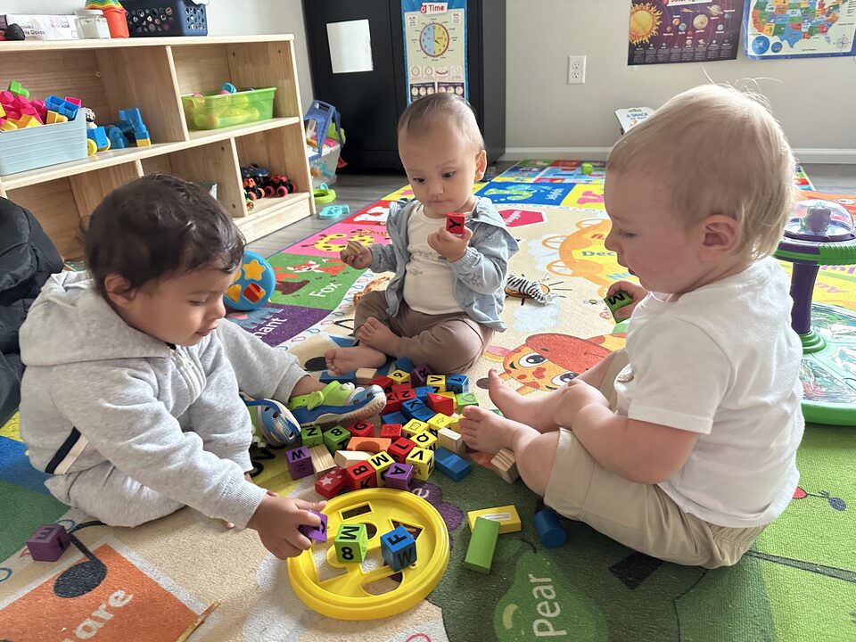 Three babies sitting together on a bright alphabet rug, turning over wooden letter blocks, with shelves of toys and learning posters behind them